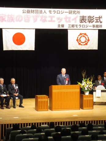 「家族のきずなエッセイ表彰式」と書かれた横断幕と国旗と三郷市旗が飾られたステージの左右に設置された椅子に胸章リボンをつけた関係者が座っているなか、演壇に立ち挨拶を行っている市長の写真