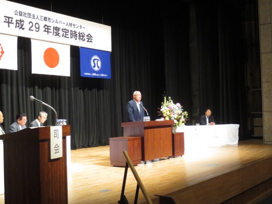 「公益財団法人三郷市シルバー人材センター平成29年度定時総会」と書かれたボードと三郷市旗・国旗・シルバー人材センターのロゴが入った旗が吊るされたステージ上で、ピンク・黄色・白などの花々で彩られた花台と並ぶ演台に立ち話をする三郷市長と、白いカバーを掛けられた席に座り話を聞く来賓者の写真