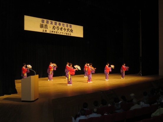 ステージ上には「演芸・カラオケ大会」と書かれた横断幕が吊るされており、紫地に白い線が入ったデザインで袖が赤色の着物を着用し金色の扇を手に持った彦川戸長生会のメンバーが、万作踊りを披露している写真