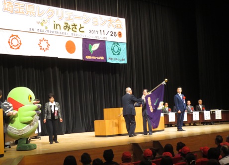 「埼玉県レクリエーション大会inみさと」と書かれた横断幕と国旗・三郷市旗を含む5つの旗がステージ上に吊るしてあり、演台の前で、紫色の県協会旗を入間市の代表の男性に手渡す三郷市長、ステージ左脇で引継ぎの様子を見守るかいちゃんと、ステージ右側に配置された席に座る来賓者、観客席に座る参加者の写真