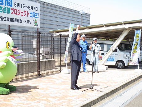 「水道週間6月1日～6月7日公園のじゃぐち点検プロジェクト出陣式！」と書かれた横断幕が固定されているフェンスの前で、右手を突き上げる三郷市長とタスキをかけたかいちゃんの写真