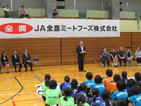 体育館に設置されたマイクスタンドの前に立ち挨拶を行っている市長の話を向かい合って体育座りをして聞いている子供たちの後ろ姿の写真