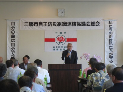 「三郷市自主防災組織連絡協議会総会」と書かれた横幕が貼られた室内で、長机が並んだ席に座っている参加者と、向かい合って演壇に立ち挨拶を行っている市長の写真