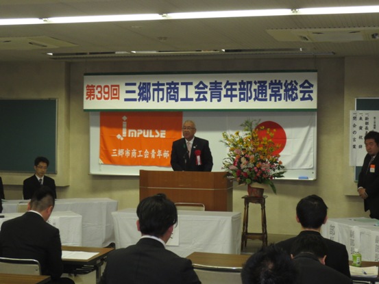 「第39回三郷市商工会青年部通常総会」と書かれたボードが天井から吊り下げられてあり、ホワイトボードには「Impulse三郷市商工会青年部」と書かれたオレンジ色の旗と国旗が貼られ、黄色・ピンク・白・緑の花々で彩られた花台と並ぶ演台に立ち祝辞を述べる三郷市長の写真