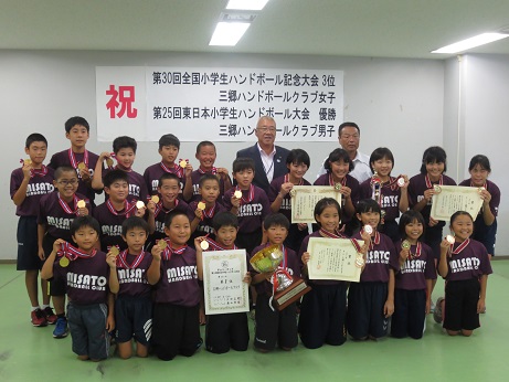 大きな優勝トロフィーを両手で持った男子選手、賞状を持った数名の選手、メダルを手に持った沢山の選手たちが三郷市長とともに並び記念撮影をしている写真