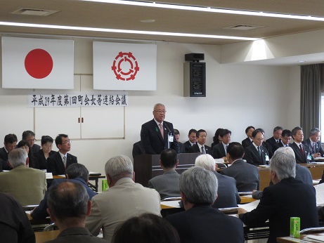 国旗と三郷市旗が天井から吊り下げられ、「平成29年度第1回町会長等連絡会議」と書かれたポスターが壁に貼られた部屋で、演台に立ち、話をする三郷市長と、席について話を聞いている参加者の写真