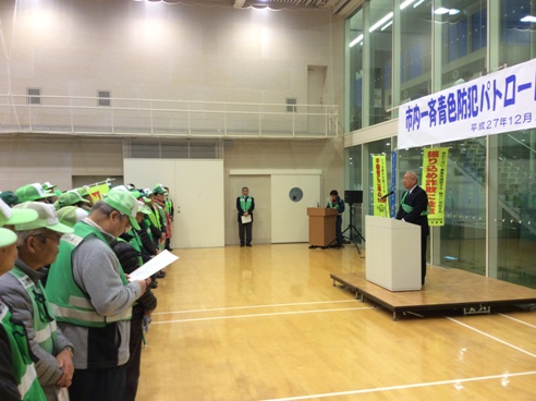 「市内一斉青色防犯パトロール出発式」と書かれた横断幕がガラス壁に貼られ、手前に設置された演壇に立ち挨拶を行っている市長の話を、向かい合って立っている緑色のキャップを被った方々が聞いている様子を右横から撮影した写真
