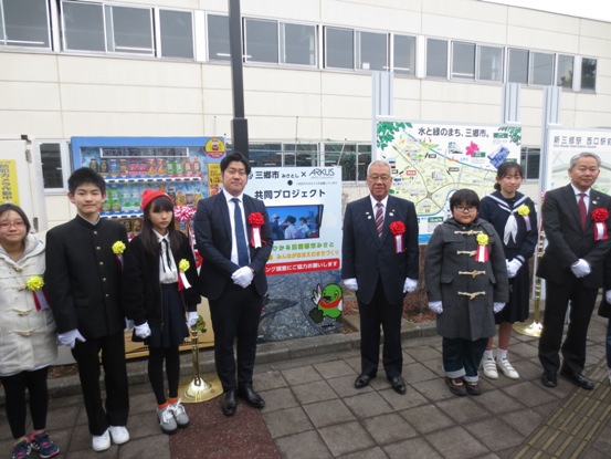 共同プロジェクトのパネルが設置された駅前の一角で、黄色や赤色の胸章をつけた関係者と一緒に記念撮影をする市長の写真