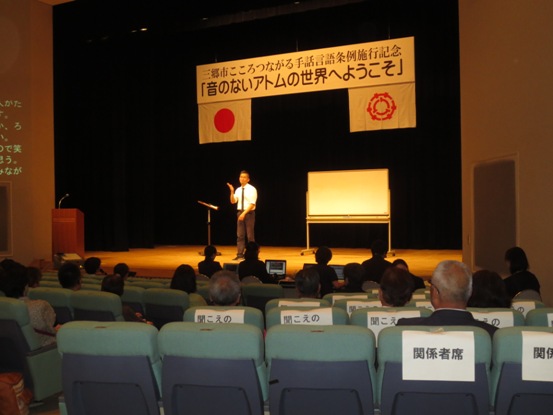 「音のないアトムの世界にようこそ」と書かれた横断幕と国旗、三郷市旗が吊り下げられているステージ上で、ホワイトボードの前に立ち手話で公演を行う砂田アトムさんと、観客席に座って公演を見ている参加者の写真