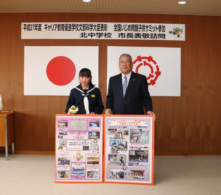 「平成27年度キャリア教育優良学校文部科学大臣表彰 全国いじめ問題子供サミット 北中学校 市長表敬訪問」と書かれた横幕と国旗と三郷市旗が飾られた室内で、2つのボードを前に立てかけた生徒会長の酒井さんと市長が並んで立っている写真