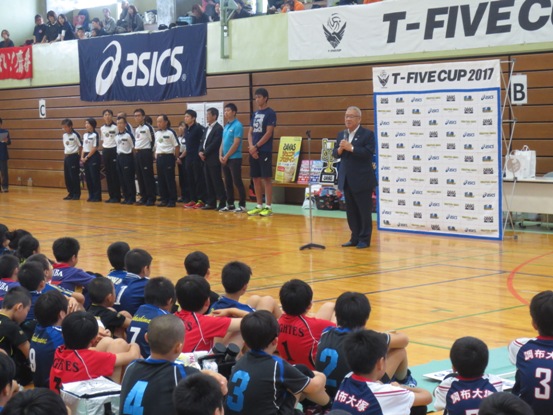 「TーFIVECUP2017」と書かれたバナースクリーンの前で、開会のあいさつの言葉を述べる三郷市長と、壁の前に横一列に並んだ関係者、体操座りで話を聞くユニフォームを着用した子どもたちの写真
