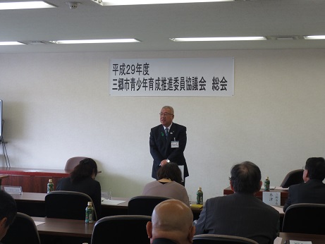 「平成29年度三郷市青少年育成推進委員協議会総会」と書かれたポスターが貼られた壁の前に立ちあいさつの言葉を述べる三郷市長と、ペットボトルのお茶が置かれた席に座って話を聞く参加者の写真
