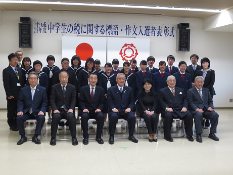 「平成29年度中学生の税に関する標語・作文入選者表彰式」と書かれた横断幕と国旗・三郷市旗が吊り下げられている壁の前で、前列に三郷市長を中央にして関係者がパイプ椅子に座り、後列に受賞したたくさんの制服姿の中学生が立っている記念写真