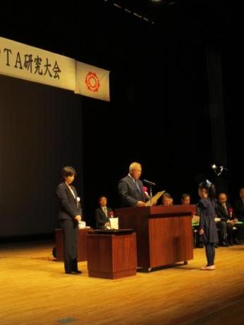 ステージの演壇を挟んで女の子と向き合って立っている市長が表彰状を読み上げている様子の写真
