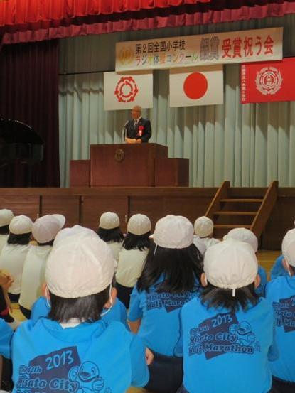 「第2回全国小学校ラジオ体操コンクール銀賞受賞を祝う会」と書かれた横断幕が飾られたステージの演壇に市長が立ち挨拶を行っているのを、赤白帽を被りお揃いの青色のシャツを着た児童達が聞いている後ろ姿の写真