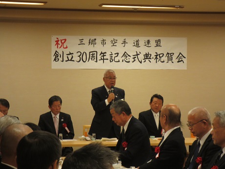 「祝三郷市空手道連盟創立30周年記念式典・祝賀会」と書かれたポスターが壁に貼り付けてあり、席を立ち挨拶の言葉を述べる三郷市長とコップの置かれた席に座り話を聞く胸章を付けた三郷市空手道連盟関係者の写真