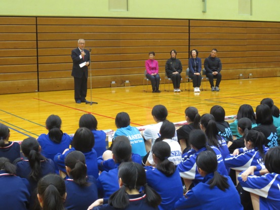 前に立ち開会の挨拶の言葉を述べる三郷市長と体操座りをし話を聞く青い体操服を着用した中学生、壁際に置かれたパイプ椅子に座る1972年ミュンヘン五輪銀メダリスト、1976年モントリオール五輪金メダリストの神白高子さん（旧姓：飯田）、1992年バルセロナ五輪銅メダリストのヨーコ ゼッターランドさん、元全日本の吉川博子さん（旧姓：津雲）さんの写真