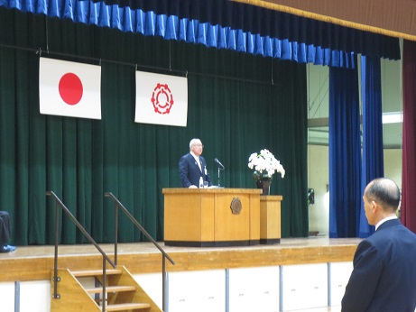 国旗と三郷市旗が吊り下げられているステージ上で、白い花が飾られた花台と並ぶ演台に立ち訓示する三郷市長の写真