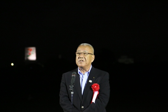 夜の屋外で、胸章を付けた三郷市長が「2017みさとサマーフェスティバル三郷花火大会」の開会セレモニーであいさつの言葉を述べている写真