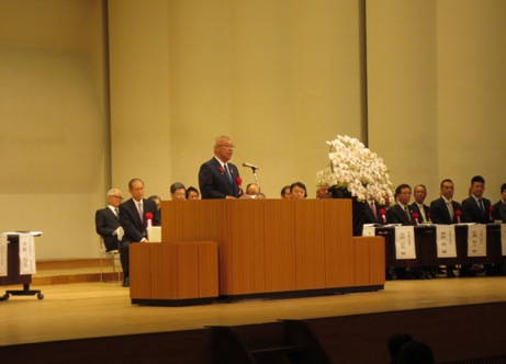 ステージの右側に設置された長机の席に関係者が座り、手前の演壇に立つ市長が挨拶を行っている写真