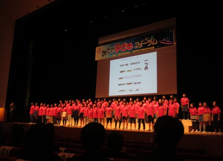 「みさと子どもフォーラム」と書かれたボードとスクリーンが吊るされているステージ上で、赤いTシャツを着用した大勢の桜小学校の子どもたちが、背後のスクリーンに映し出された活動内容についての発表を行っている写真