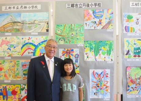 学校別に掲示ボードに展示されている絵画の作品の前で、グレー色のシャツを着た女の子と市長が横に並んで立っている記念写真