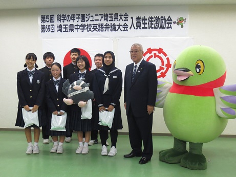 「第5回科学の甲子園ジュニア埼玉県大会第69回埼玉県中学校英語弁論大会入賞生徒激励会」と書かれた横断幕と国旗・三郷市旗が貼り付けられた壁の前で、中央でつぶちゃんのぬいぐるみを両手に持った女子学生と前後2列に並んだ白い袋を手に持った合計7名の入賞者たちが傍らに立つ三郷市長とかいちゃんとともに記念撮影をしている写真