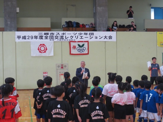 「三郷市スポーツ少年団平成29年度全団交流レクリエーション大会」と書かれた横断幕と三郷市旗、重なった5つの輪の上に双葉マークが描かれているJSBの旗が貼られた壁を背に、開会の言葉を述べる三郷市長と、チームごとに整列し立って話を聞く子どもたちの写真