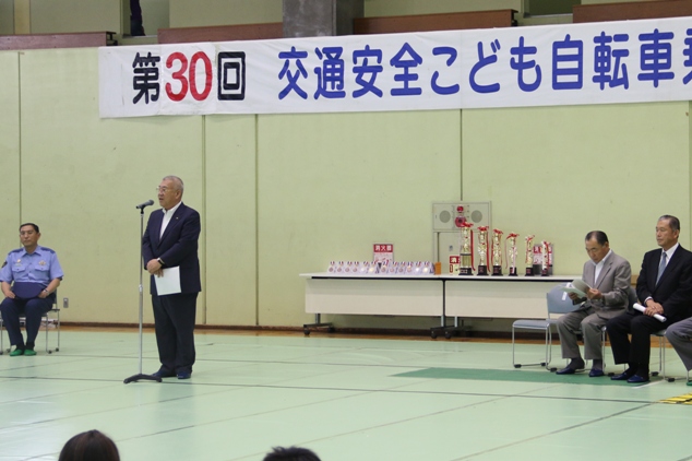 「第30回こども自転車乗り大会」と書かれた横断幕が貼られた会場のマイクスタンドの前で挨拶を行っている市長の写真