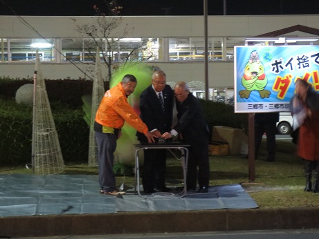 折り畳み式の小さなテーブルの上に置いたボタンを、2名の男性と市長が一緒に押そうとしている点灯の瞬間の様子の写真