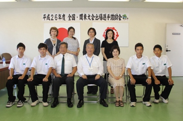 平成26年度全国・関東大会出場選手激励会と書かれた横断幕、国旗と三郷市旗が飾られた室内で水泳で優秀な成績を収めた早稲田中学校の4名の男子生徒と男性と女性の教師が市長を挟んで横一列に並んで椅子に座り、後方に4名の女性が立っている記念写真