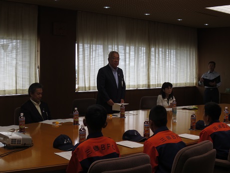 席に立ち祝辞を述べる三郷市長と、ペットボトルのお茶と資料が置かれている席に座り話を聞く関係者、市長の対面に座ったオレンジとネイビーの制服を着用した少年消防クラブのメンバーたちの写真