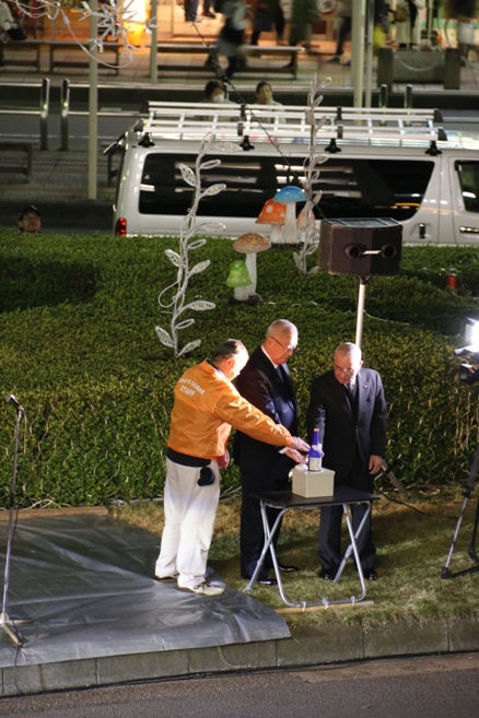 三郷市長と関係者の男性2名が、JR三郷駅南口の芝生の上で黒い机の上に置かれている青い酒瓶の後ろに設置された白いボタンを押しているイルミネーション点灯式の様子の写真