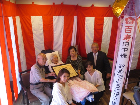 割られたくす玉の中に「祝・百寿 田中様おめでとうございます」と書かれたリボンが掛けてあり、三郷市長が田中たけさんと表彰状を持つご家族とともに紅白幕が掛けられた部屋で記念撮影をしている写真