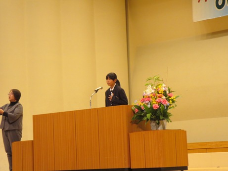 ステージの左側に立つ手話通訳者の女性が手話を行いながら、檀上に立つ女性生徒が話をしている青少年の主張大会の様子の写真