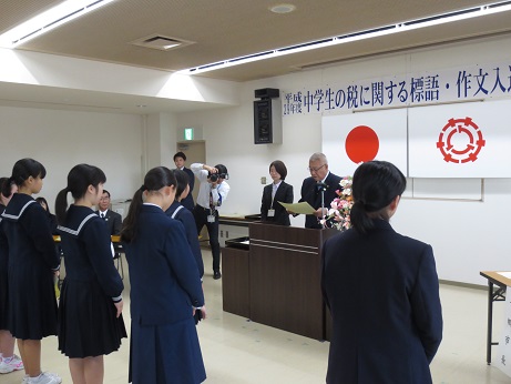 横断幕と国旗・三郷市旗が吊り下げられている壁の前で、演台に立ち表彰状の内容を読み上げる三郷市長と、市長と向かい合って立ち、話を聞く表彰された中学生たちの写真