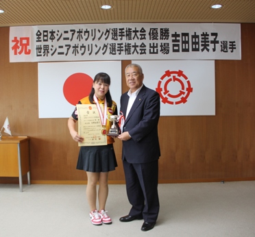 「祝 全日本シニアボウリング選手権大会優勝・世界シニアボウリング選手権大会出場 吉田由美子選手」と書かれた横幕が飾られた室内で、首にメダルをかけた吉田選手とトロフィーを持った市長が並んで立っている記念写真