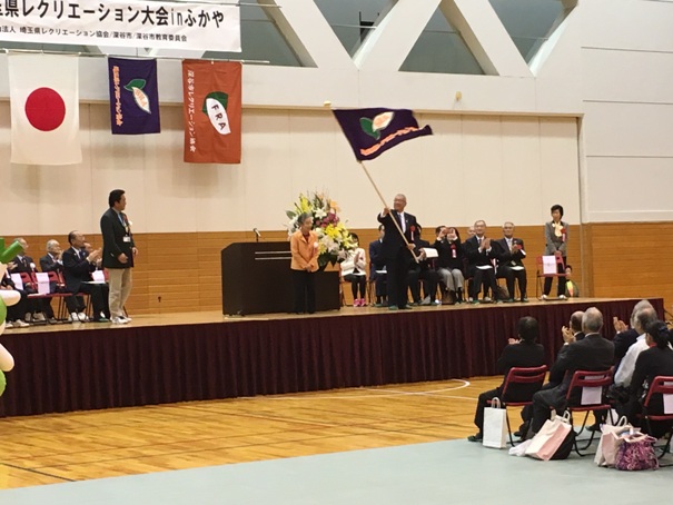 「埼玉県レクリエーション大会inふかや」と書かれた横断幕が貼られた会場のステージ左右に胸章リボンをつけた関係者が着席して拍手を送るなか、演壇の前にオレンジ色のジャケットを着用した女性が立ち、右側の市長が大きな旗を受け取った様子の写真