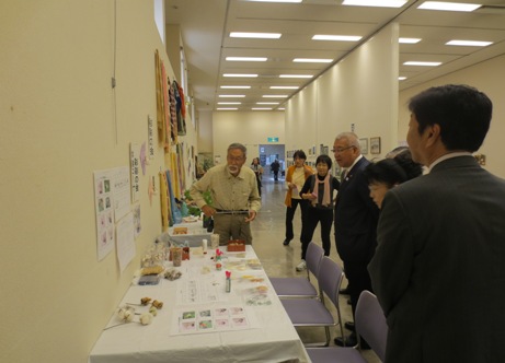 白色のクロスがかけられたテーブルに作品が展示されている傍に立つ出展者の男性に、市長や3名の関係者が並んで説明を受けている様子の写真