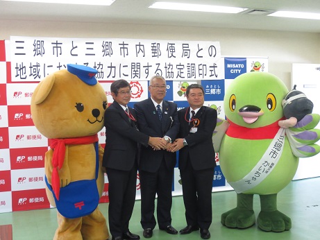 「三郷市と三郷市内郵便局との地域における協力に関する協定調印式」と書かれたポスターが貼りつけられている三郷市と郵便局のバナースタンドの前で、手を合わせて立つ三郷市長と、2名の郵便局員を、かいちゃんとぽすくまが間に挟み記念撮影をしている写真