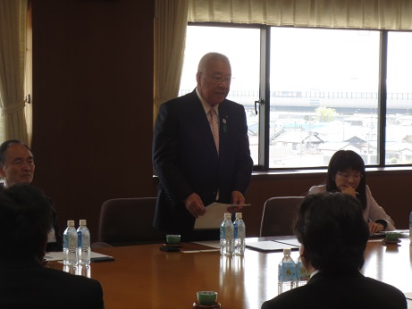資料を持ちながら立ってあいさつの言葉を述べる三郷市長と、ペットボトルの水が2本ずつ置かれた席に座り話を聞く参加者の写真