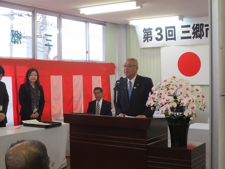 「第3回三郷市景観賞表彰式」と書かれた横断幕と国旗・三郷市旗が貼り付けられている壁の前で、白色の花々で彩られた花台と並ぶ演台に立ち挨拶の言葉を述べる三郷市長と、窓際に掛けられた紅白幕の前に立って話を聞く女性の写真