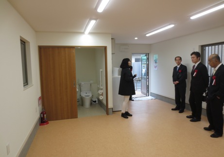 戸が開かれたトイレの近くに赤の胸章を付けた3名の男性と女性が立ち、いずみふれあい集会所を内覧している様子の写真