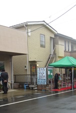 ベージュ色の2階建ての建物で、一階部分にガラスドアがあり、外側に2階に続く階段があるいずみふれあい集会所と、建物の前に設置された緑色のバザーテントの下にレッドカーペットが敷かれている写真
