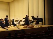 ステージの後方に座っている生徒たちの演奏に合わせて、3名の女子生徒がそれぞれの楽器を持ちステージの前方に間隔をあけて立ってソロパートの演奏を行っている様子を左斜めから撮影した写真
