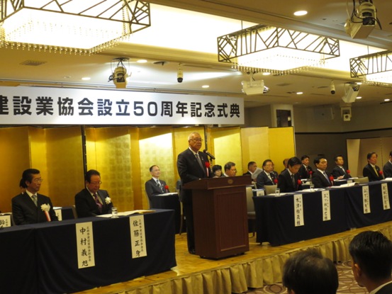 「三郷市建設業協会設立50周年記念式典」と書かれたボートが天井から吊り下げられ、その下には金色のジャバラの屏風が置かれたステージの上で、演台の前に立ち祝辞の言葉を述べる三郷市長と、その左右で前後に並んだ紺のカバーが掛けられ席札が貼られた席に座り話を聞く胸章を付けた来賓者の写真