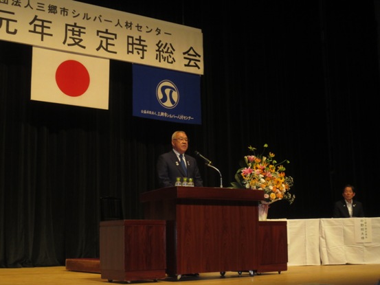 横断幕と2枚の旗が飾られたステージの右側の席に関係者が座り、演題で挨拶を行っている市長の写真
