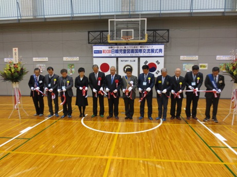 「第30回日韓児童図画国際交流展式典」と書かれた横断幕の前で関係者が横一列に並びテープカットを行っている様子の写真
