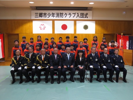 三郷市旗・国旗・消防旗の上に「三郷市少年消防クラブ入団式」と書かれた横断幕が吊り下げられている壁の前で、三郷市長と関係者が制服を着用した入団者22名とともに記念撮影をしている写真