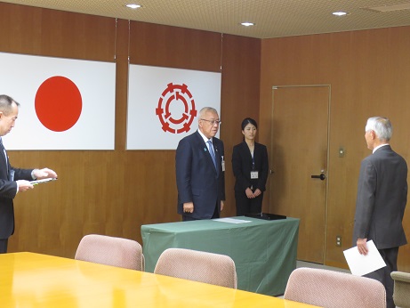 国旗と三郷市旗が吊り下げられている壁の前で、緑のカバーが掛けられ黒い賞状盆が置かれた台の後ろに立ち挨拶の言葉を述べる三郷市長と向かい合って立つ公平委員会委員の男性の写真